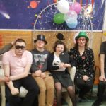 Grupa siedzących osób w karnawałowych strojach, w śmiesznych okularach i kolorowych kapeluszach.