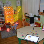 Trzech chłopców siedzi na dywanie, jeden przy stole i bawi sie kolorowymi światłami. W tle kolorowe świecące kolumny.