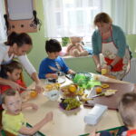 Pięciu małych chłopców siedzi przy stole. Dwie kobiety kroją owoce. Na stole owoce, miski, deski do krojenia, talerze.