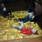 Jeden chłopiec leży na kolanach kobiety a dwóch na podłodze. Podłoga podświetlona jest widokiem morskich kamieni.